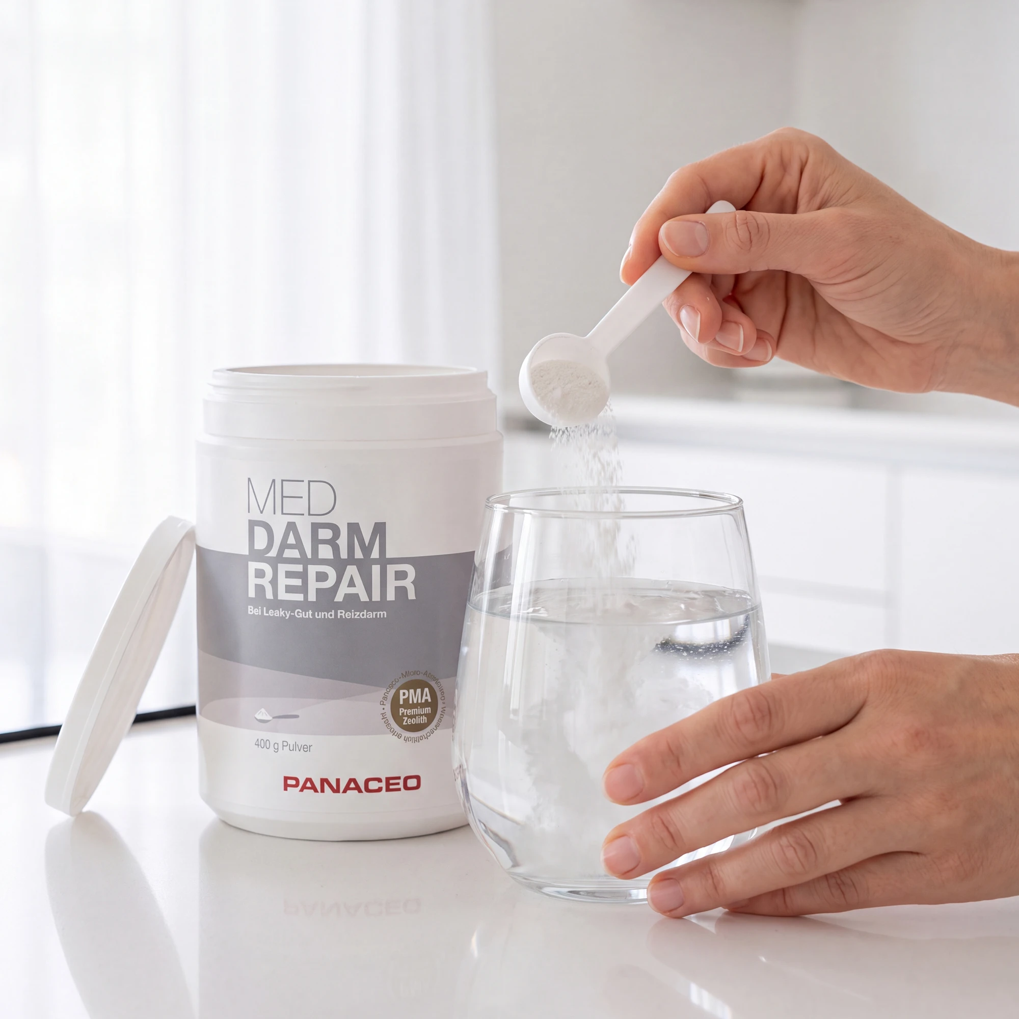 PANACEO MED DARM REPAIR Pulver 400 g – Hand dosiert Pulver mit Messlöffel in ein Glas Wasser neben geöffnetem Produktbehälter