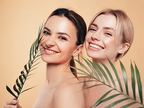 Zwei lächelnde Frauen mit gesunder Haut stehen vor beigem Hintergrund und halten grüne Palmblätter. Das Bild symbolisiert natürliche Schönheit, Pflege und Hautgesundheit mit PMA-Zeolith.