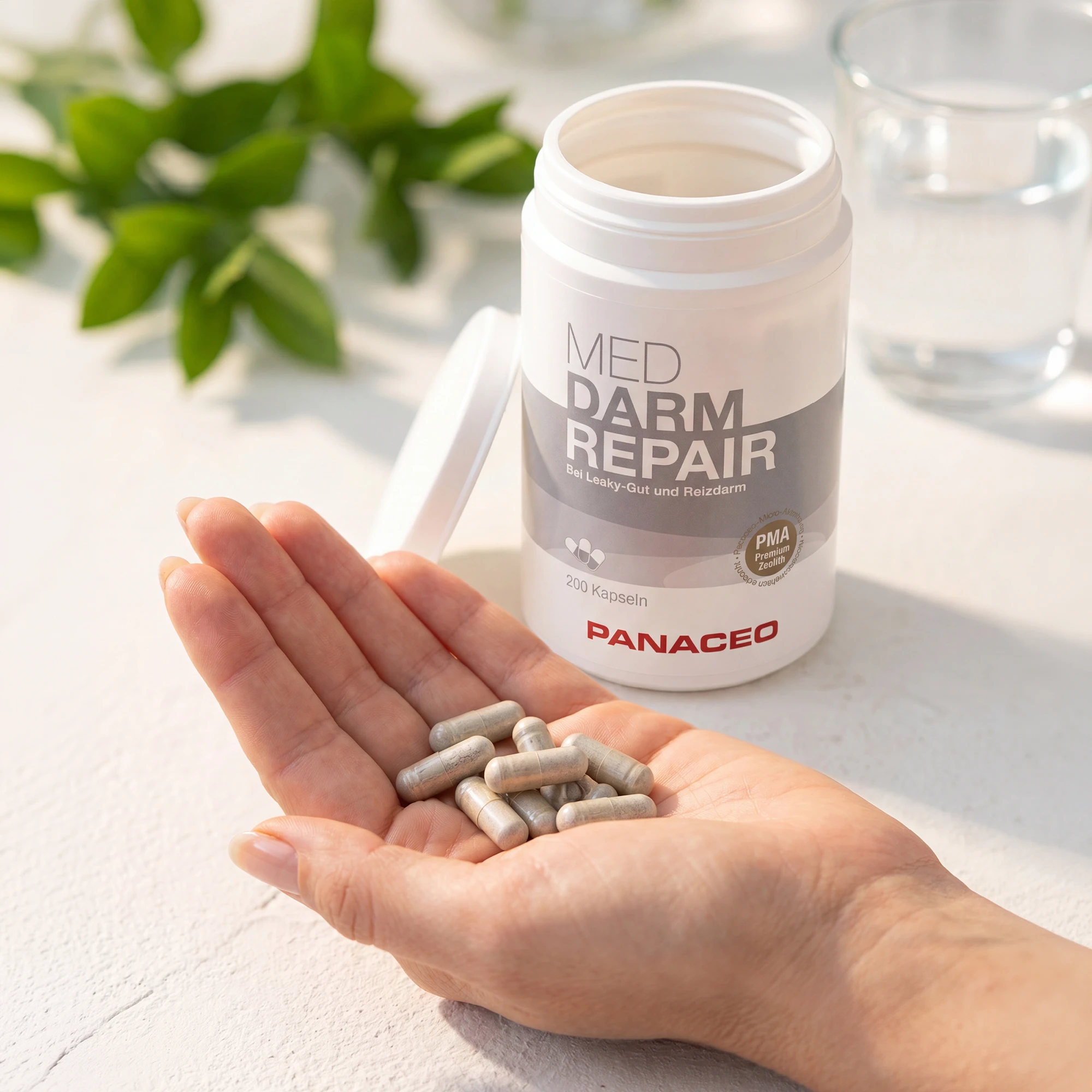 Offene Dose PANACEO MED DARM REPAIR Kapseln 200 Stk. mit Kapseln in einer Hand, daneben ein Glas Wasser